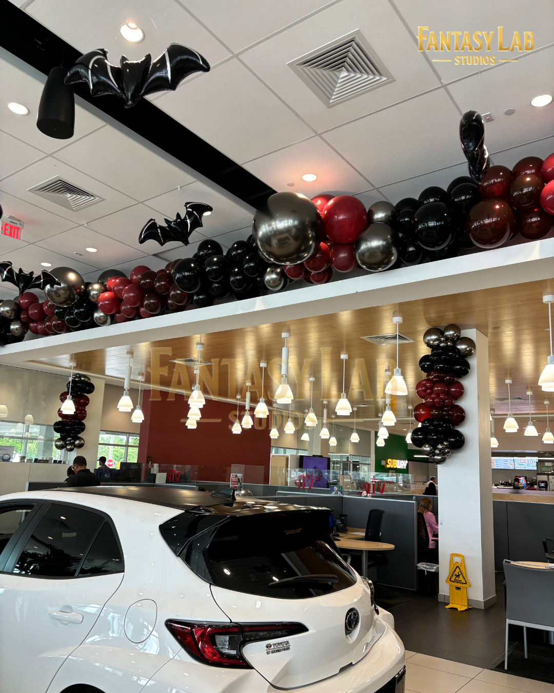 Ceiling balloon garland with bat silhouettes spanning a modern auto showroom.
